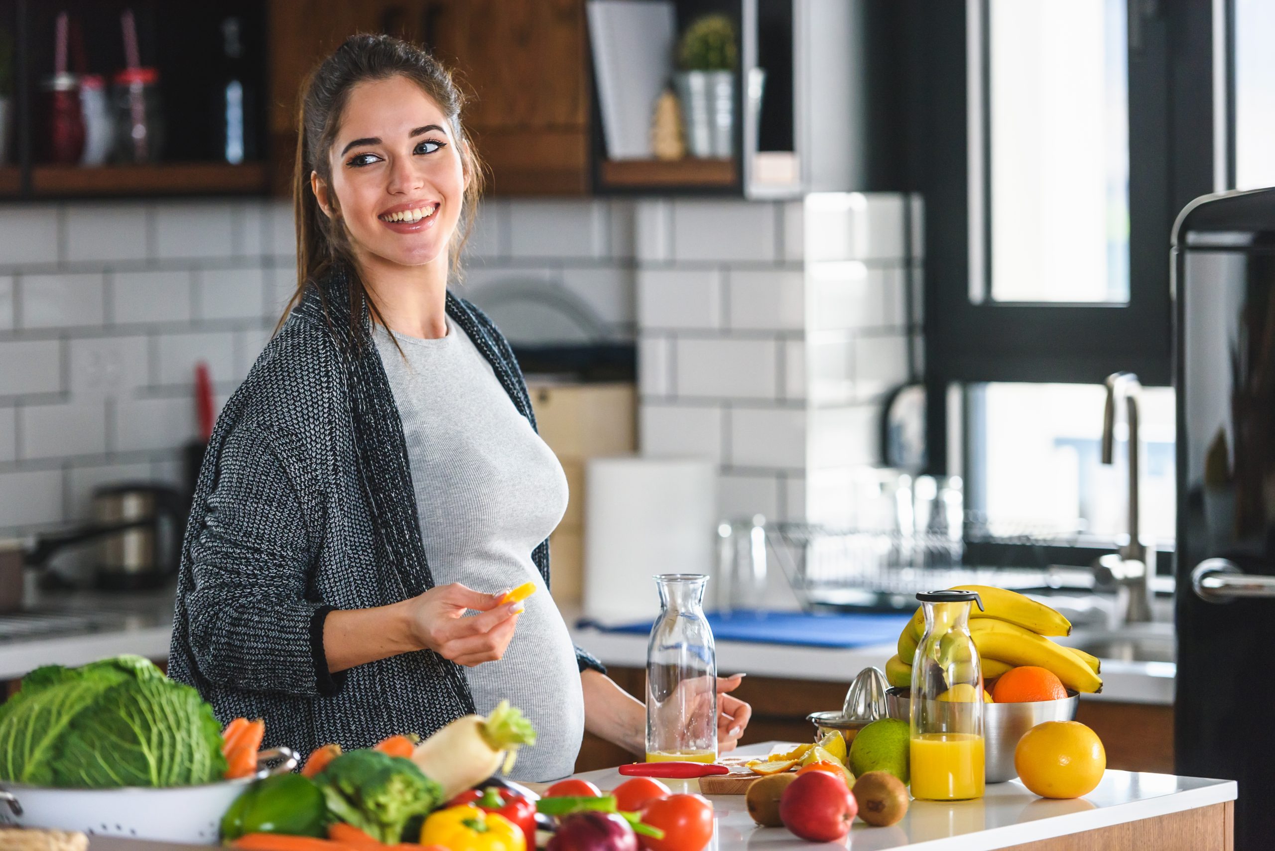 pregnant woman standing in kitchen with lots of fruit and vegetables in front of her